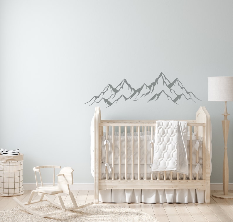 May include: A white crib with a white quilt and a gray mountain range wall decal in a nursery setting.