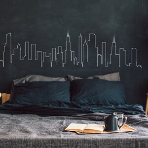 K&ouml;nnte beinhalten: Ein Schlafzimmer mit einer dunkelgrauen Wand, auf der eine wei&szlig;e Umrisszeichnung einer Skyline zu sehen ist. Das Bett hat eine graue Bettdecke und dunkelblaue Kissen. Ein kleiner Holz-Beistelltisch mit einer Kerze und einer Topfpflanze steht neben dem Bett.