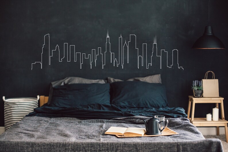 K&ouml;nnte beinhalten: Ein Schlafzimmer mit einer dunkelgrauen Wand, auf der eine wei&szlig;e Umrisszeichnung einer Skyline zu sehen ist. Das Bett hat eine graue Bettdecke und dunkelblaue Kissen. Ein kleiner Holz-Beistelltisch mit einer Kerze und einer Topfpflanze steht neben dem Bett.