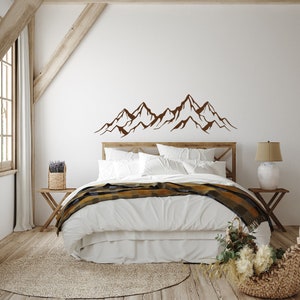 May include: A white bed with a brown blanket and a mountain range wall decal above the headboard. The bed is in a room with a wooden floor and a wooden side table with a vase of flowers.