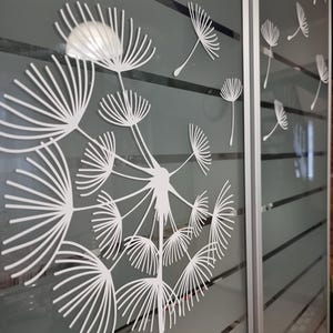 May include: A white dandelion design on a frosted glass sliding door. The dandelion is in the center of the image and has many seeds blowing in the wind. The door is made of frosted glass and has a silver frame.