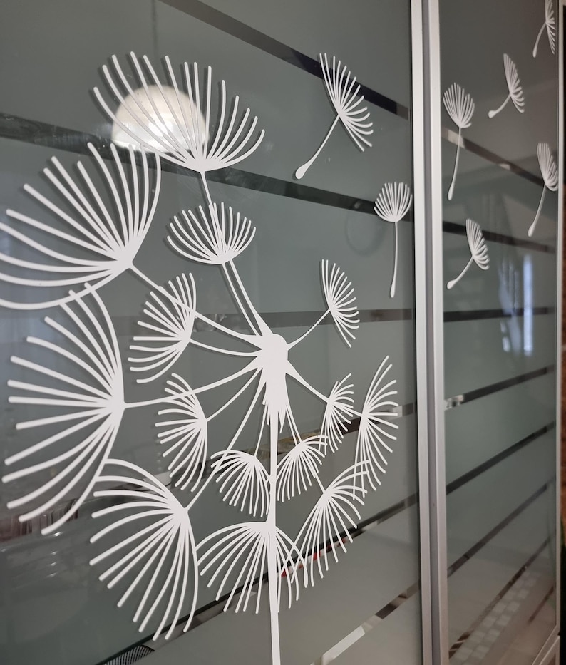 May include: A white dandelion design on a frosted glass sliding door. The dandelion is in the center of the image and has many seeds blowing in the wind. The door is made of frosted glass and has a silver frame.