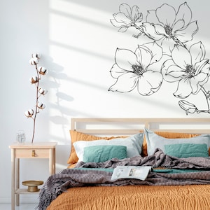 May include: A bedroom with a bed with a brown and white striped blanket and a teal and white striped pillow. There is a white wall with a black floral decal on it. There is a small wooden nightstand with a glass vase and a gold tray on it.
