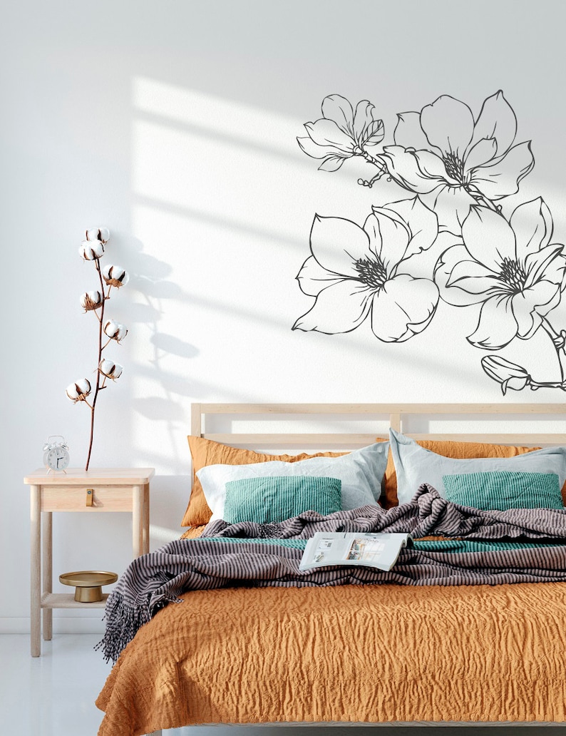 May include: A bedroom with a bed with a brown and white striped blanket and a teal and white striped pillow. There is a white wall with a black floral decal on it. There is a small wooden nightstand with a glass vase and a gold tray on it.