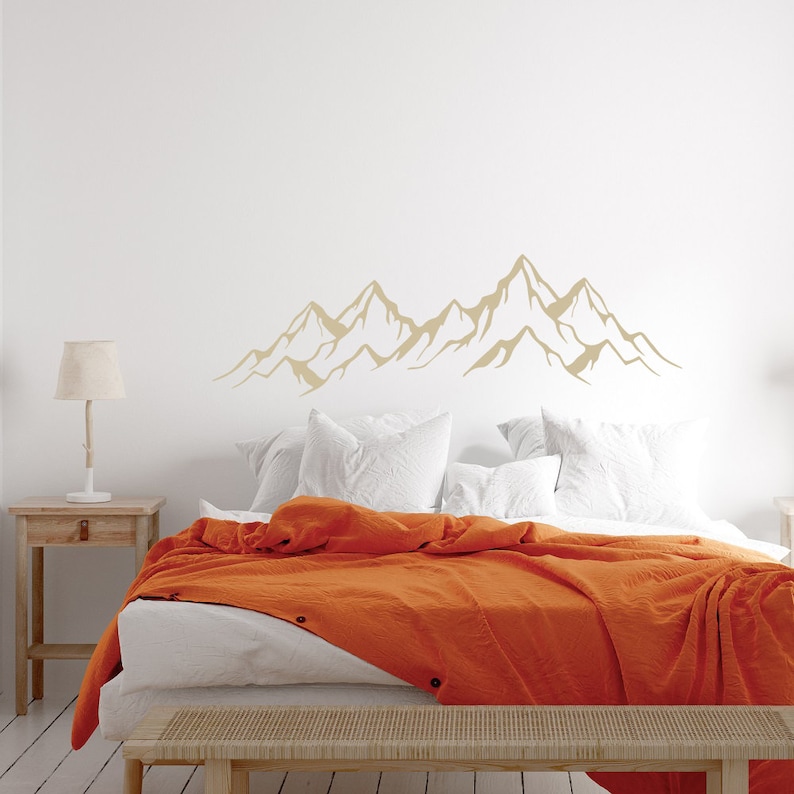 May include: A white bed with an orange blanket and two white pillows. A mountain range silhouette decal is on the wall behind the bed.