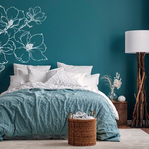 May include: A teal bedroom with a bed with white bedding and a blue textured throw blanket. A white floral design is on the wall behind the bed. A wicker basket is at the foot of the bed. A floor lamp with a white shade is in the corner of the room.