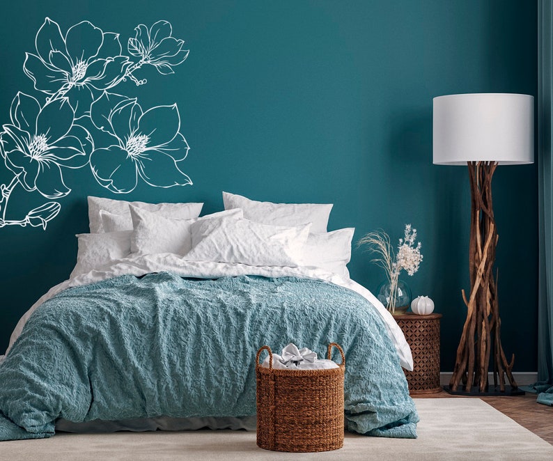 May include: A teal bedroom with a bed with white bedding and a blue textured throw blanket. A white floral design is on the wall behind the bed. A wicker basket is at the foot of the bed. A floor lamp with a white shade is in the corner of the room.