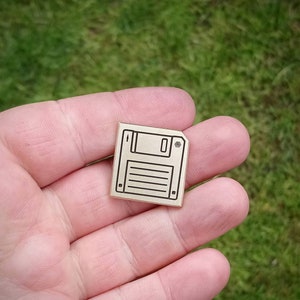 May include: A gold-colored metal pin shaped like a floppy disk. The pin has a black outline and a small square hole in the top right corner.