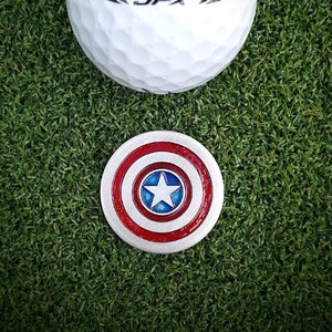May include: A white golf ball with the text "<JPX>" printed on it, and a red, white and blue golf ball marker with a star in the centre.