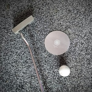 May include: A silver putter with a black grip, a white golf ball, and a silver disc with the text "Never Stop" on a grey carpet.
