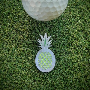 May include: A silver pineapple golf ball marker with a green pineapple design. The marker is on a green artificial grass surface.