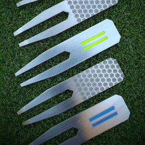 Pode incluir: Cinco ferramentas de reparo de divots de golfe prateadas com diferentes acentos coloridos. As ferramentas têm um design de garfo com uma superfície texturizada. Os acentos são vermelho, cinza, verde, cinza e azul.