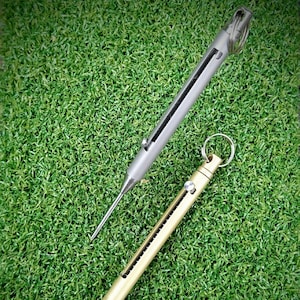 Puede incluir: Dos herramientas metálicas con llaveros descansan sobre una superficie verde. Una es plateada, la otra dorada. Ambas tienen una serie de pequeñas aberturas a lo largo del lateral. La herramienta plateada tiene un extremo largo, fino y puntiagudo. La herramienta dorada tiene un extremo redondeado.