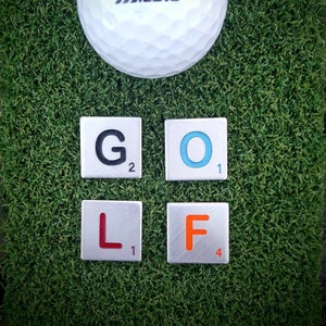 以下が含まれることがあります： 白いミズノゴルフボール、数字の3と黒いロゴが、"GOLF"と綴られた4つのスクラブルタイルの上にあります。タイルには、緑色の表面に黒、青、赤、オレンジの文字があります。