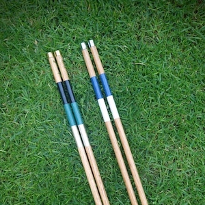 Puede incluir: Cuatro mazos de croquet de madera con bandas de colores negro, verde, blanco y azul. Los mazos descansan sobre hierba verde, lo que sugiere un juego al aire libre. Los mazos tienen puntas metálicas.
