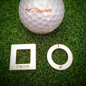 May include: A white golf ball with the word "Mizuno" printed on it, a gold square with the word "FORSYTH" engraved on it, and a gold ring with the letters "RF" engraved on it. All items are on a green artificial grass surface.