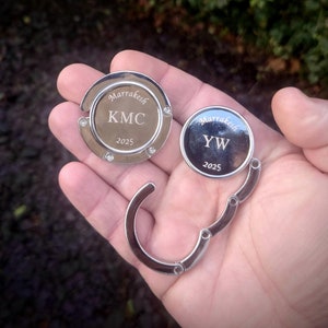 Könnte beinhalten: Zwei silberfarbene Taschenclips mit eingraviertem Text. Der erste Clip sagt "Marrakesh KMC 2025" und der zweite Clip sagt "Marrakesh YW 2025".