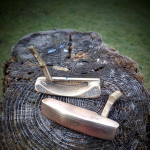 Könnte beinhalten: Zwei vintage goldfarbene Golf-Putter, einer mit dem Wort "PING" und der andere mit dem Wort "ZING" an der Seite. Die Putter liegen auf einer Holzoberfläche.