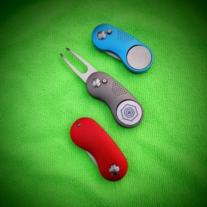 Puede incluir: Tres herramientas para reparar divots de golf en azul, gris y rojo, dispuestas sobre una superficie verde. Cada herramienta tiene un color y diseño distintos, con un marcador de bola magnético. Las herramientas están diseñadas para reparar divots en un campo de golf.