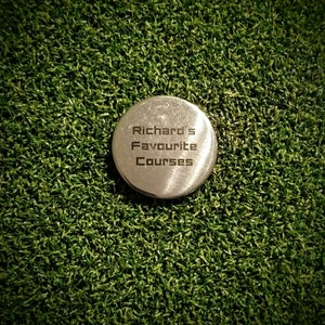 May include: A silver metal golf ball marker with the text "Richard's Favourite Courses" engraved on it. The marker is sitting on a green artificial grass surface.