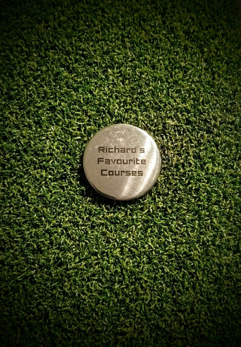 May include: A silver metal golf ball marker with the text "Richard's Favourite Courses" engraved on it. The marker is sitting on a green artificial grass surface.