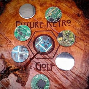 May include: Eight round, silver-colored, circuit board-style golf ball markers with the text "Future Retro Golf" on a wooden surface.
