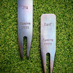 May include: Two rectangular metal golf divot tools with a dark, iridescent finish. The left tool is engraved with JYA, BANDON, and 2022. The right tool is engraved with DAVE, BANDON, and 2022. Both feature a two-pronged bottom for turf repair.