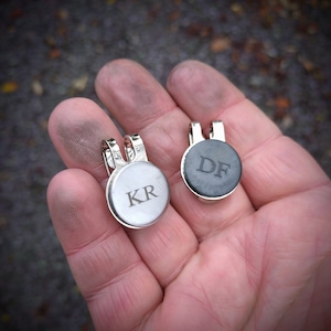 Peut inclure: Deux marqueurs de balle de golf personnalisés, un argenté avec les initiales "KR" et un noir avec les initiales "DF".