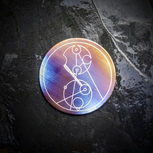 May include: A circular metal disc featuring an iridescent finish that transitions from deep blue to warm gold. The surface is engraved with a white, intricate geometric pattern consisting of interconnected circles, lines, and small dots.