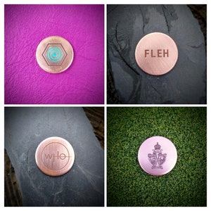 Może przedstawiać: Cztery różne znaczniki do piłek golfowych z miedzi o różnych wzorach. Pierwszy znacznik ma geometryczny wzór w kolorze turkusowym i brązowym. Drugi znacznik ma wygrawerowane słowo "FLEH". Trzeci znacznik ma wygrawerowane słowo "WHO". Czwarty znacznik ma herb z koroną i dwoma tarczami.