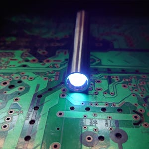 Pu&ograve; includere: Primo piano di un tubo metallico argentato che emette una luce bianca brillante. Il tubo &egrave; posizionato verticalmente sopra un circuito stampato verde con componenti elettronici.
