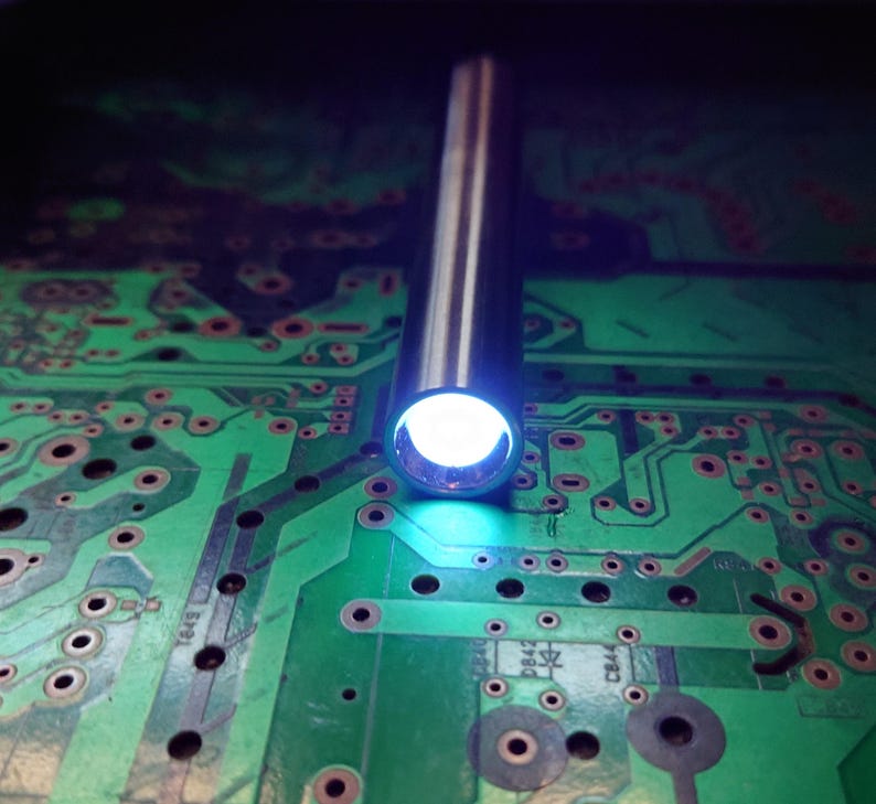 Pu&ograve; includere: Primo piano di un tubo metallico argentato che emette una luce bianca brillante. Il tubo &egrave; posizionato verticalmente sopra un circuito stampato verde con componenti elettronici.