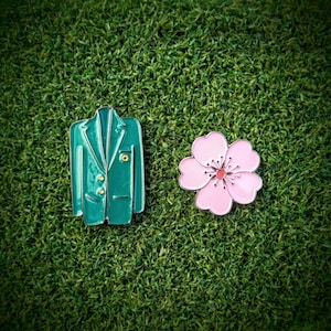 May include: A green enamel pin shaped like a blazer and a pink enamel pin shaped like a flower on a green artificial grass background.