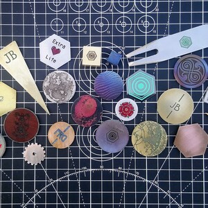 May include: A collection of small, round and hexagonal metal tokens with various designs and colours, including a gold token with the text "Future Retro Gear" and a gold token with the text "Extra Life".