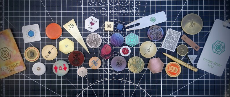May include: A collection of small, round and hexagonal metal tokens with various designs and colours, including a gold token with the text "Future Retro Gear" and a gold token with the text "Extra Life".