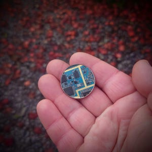 May include: A close-up of a small, round circuit board with blue and gold components. The board is held in a hand, with a blurred background of red and brown.