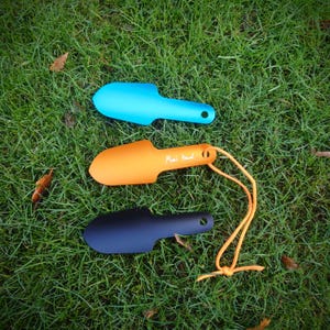 Peut inclure: Trois petites truelles de jardin, bleues, orange et noires, posées sur de l'herbe verte. La truelle orange porte l'inscription "Mimi's travel" et une corde orange. Chaque truelle a un trou dans le manche.