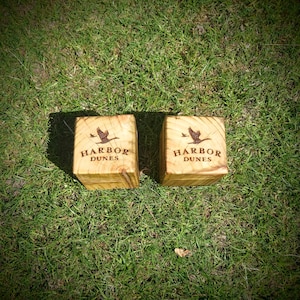 May include: Two light brown wooden cubes featuring a dark brown engraved graphic of a flying bird above the text HARBOR DUNES. The wood grain is visible on the top surfaces of both square blocks, which are positioned side by side on green grass.