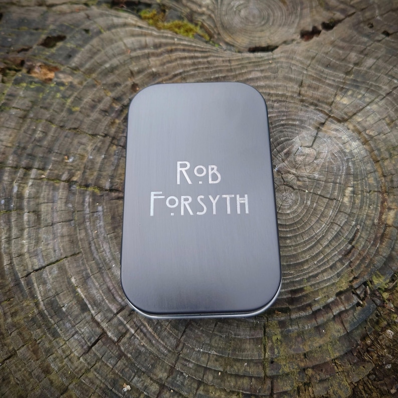 May include: A silver metal tin with the text "Rob Forsyth" engraved on the front.