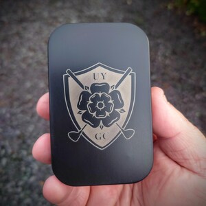 May include: Black metal tin with a silver engraved logo. The logo features a shield with a rose and crossed golf clubs. The text "UY GC" is engraved below the rose.