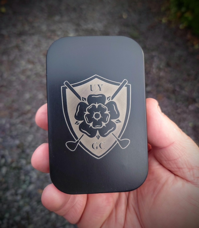 May include: Black metal tin with a silver engraved logo. The logo features a shield with a rose and crossed golf clubs. The text "UY GC" is engraved below the rose.