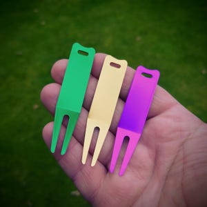 Op de afbeelding: Drie golfdivot-reparatietools in groen, goud en paars. Elke tool heeft een rechthoekige bovenkant met een gat en een vork met twee tanden aan de onderkant. De tools worden in een hand gehouden tegen een vage groene achtergrond.