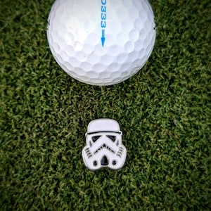 May include: A white golf ball with the text "EAD333" printed on it. A white and black Stormtrooper helmet golf ball marker is on the green grass.