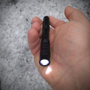 May include: A small, black flashlight with a textured body and a bright white light beam.