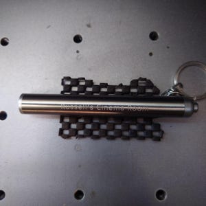 Pu&ograve; includere: Un portachiavi in metallo color argento con la scritta incisa "Russell's Cinema Room". Il portachiavi include una torcia cilindrica e un elemento di design nero a scacchi. &Egrave; presente un portachiavi argentato.