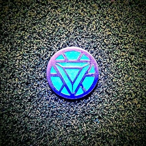 Puede incluir: Un pin de metal azul y plateado con un diseño estilizado de un círculo con un triángulo en su interior. El pin está sobre un fondo verde.