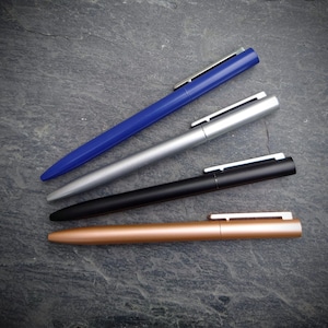 May include: Four pens in different colors: blue, silver, black, and copper. The pens are laid out in a row on a gray surface.