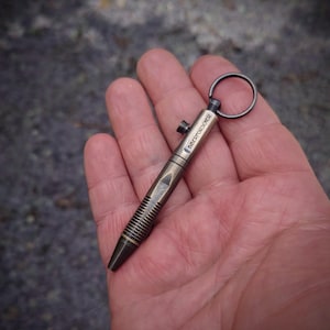 May include: A small, metallic, bronze-toned pen with a textured, ribbed grip section and a smooth barrel. The barrel features the engraved text BIDERFORSCHER and includes a small side-mounted bolt mechanism and a circular metal ring at the top end.