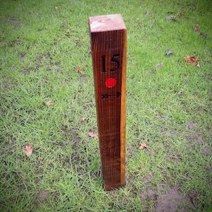 May include: A wooden post with the number 15 painted on it, a red dot, and an arrow pointing to the right. The post is standing in a grassy area.