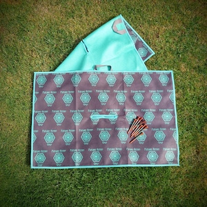 May include: A teal and grey golf mat featuring a repeating geometric design and the text "Future Retro" and "Golf". The mat is folded and has a handle. A bundle of golf tees is on the mat.